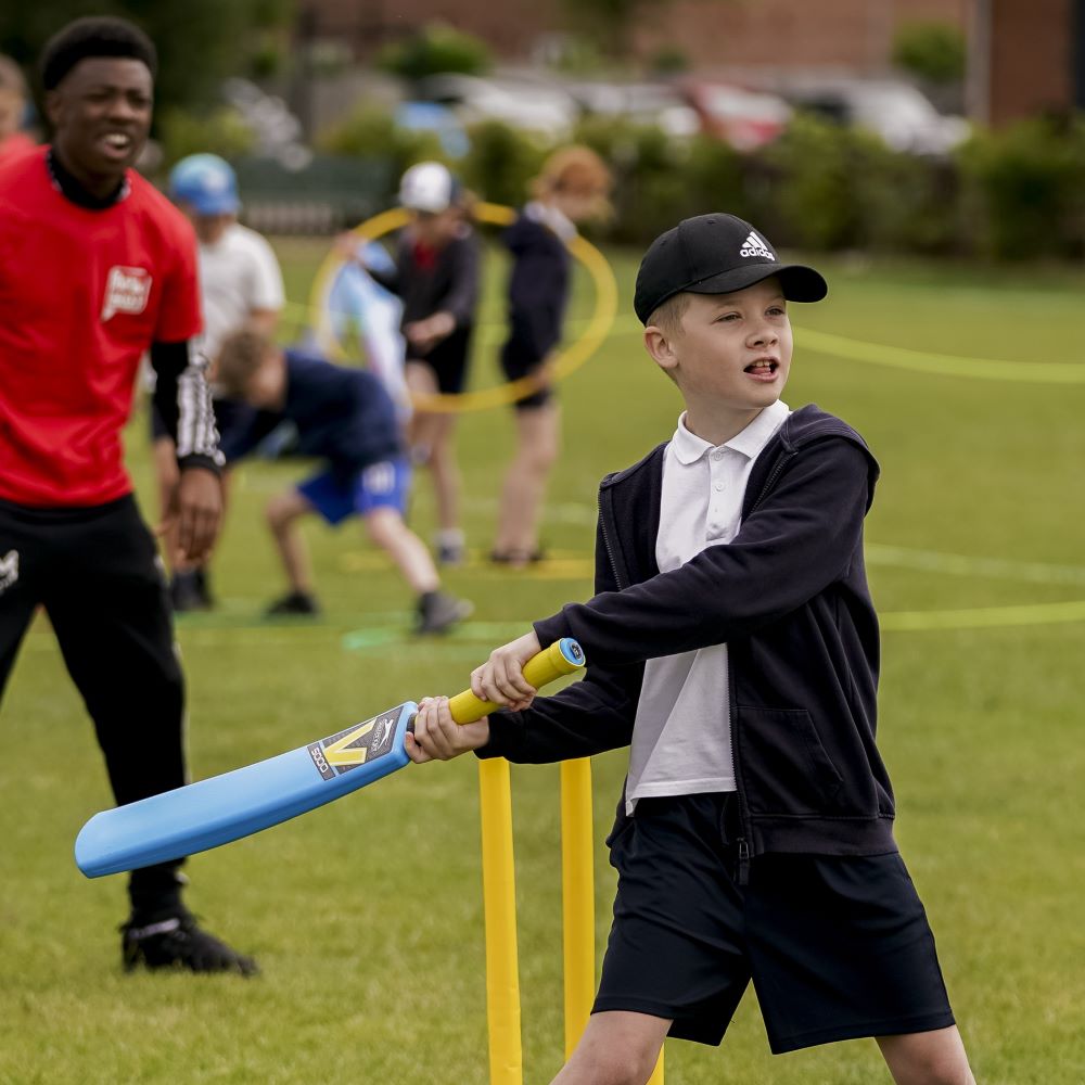 Boy in cap and PE kit swinging a blue cricket bat