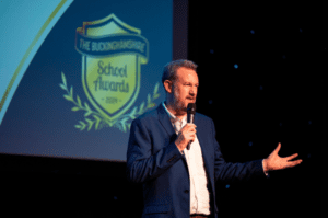 Comedian Alex Horne speaking on stage in front of Buckinghamshire School Awards banner