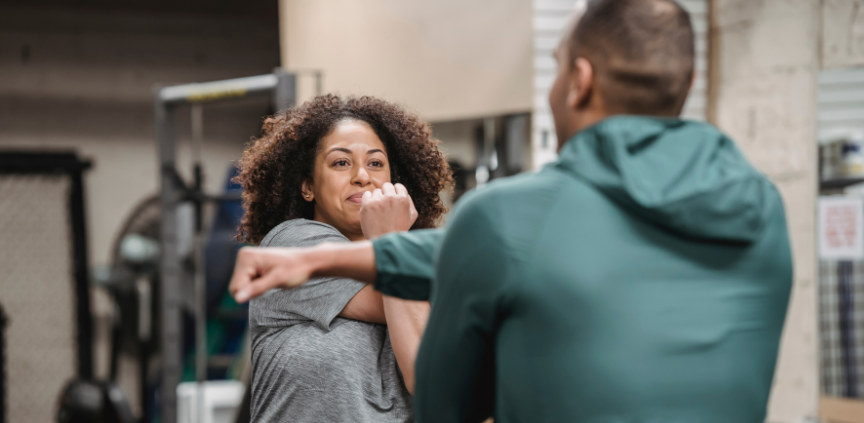 Two individuals in a gym setting, one person stretching by pulling an arm across the chest while the other, facing away from the camera, appears to assist or instruct. Gym equipment like weights and machines are visible in the background