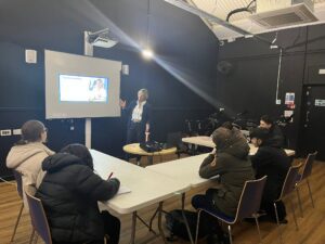 Young people sat in a classroom setting, looking at a screen whilst an adult is presenting 