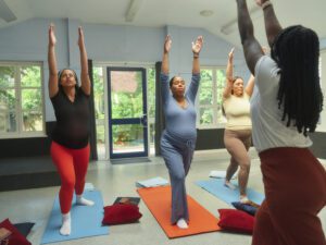 Three pregnant women stretching on mats, standing in a pose with their arms straight up, above their head. In front of them is a female instuctor.